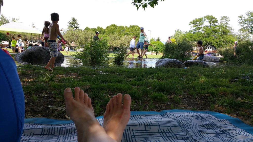 A view of a woman's feet in a shaded area as she looks on to her children playing in the water.