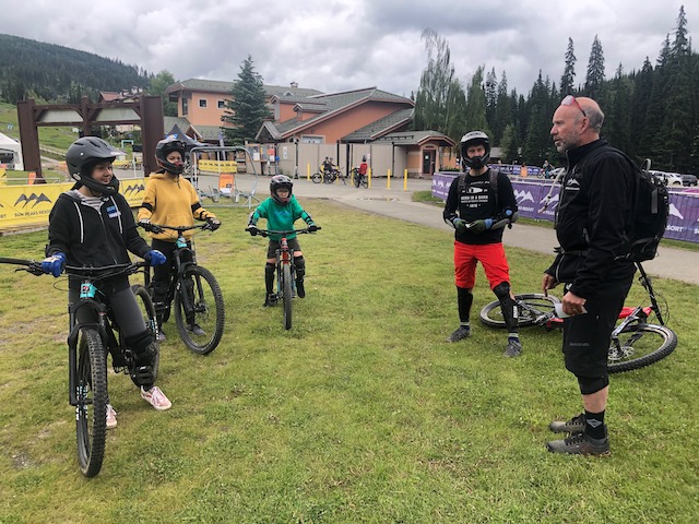 Three kids stand on downhill mountain bikes with their father as they listen to their instructor during their lesson.