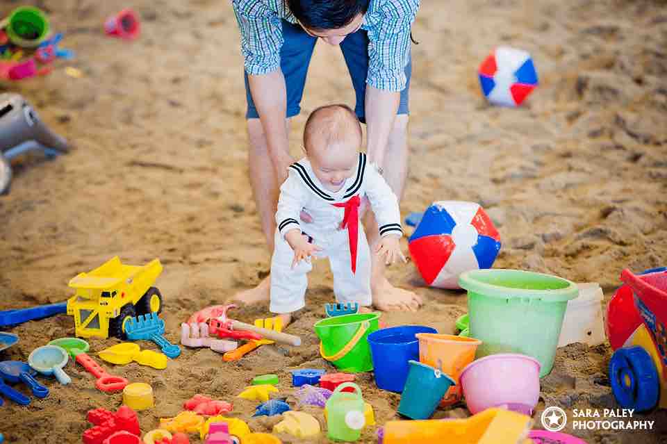 A dad holds a small toddler, dressed in a sailor suit, around the waist as he reaches for a green sand bucket at 6Pack Beach.