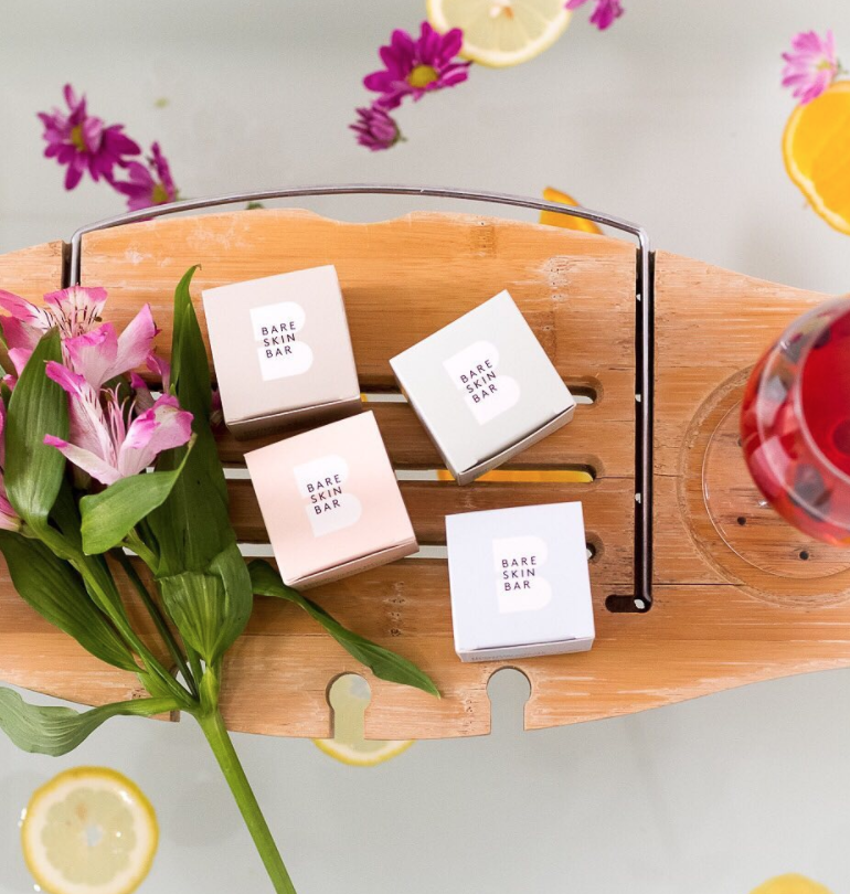 A photo of four boxes of Bare Skin Bar soaps in pink, green, brown and light blue boxes sitting on a wood tray next to fresh flowers.