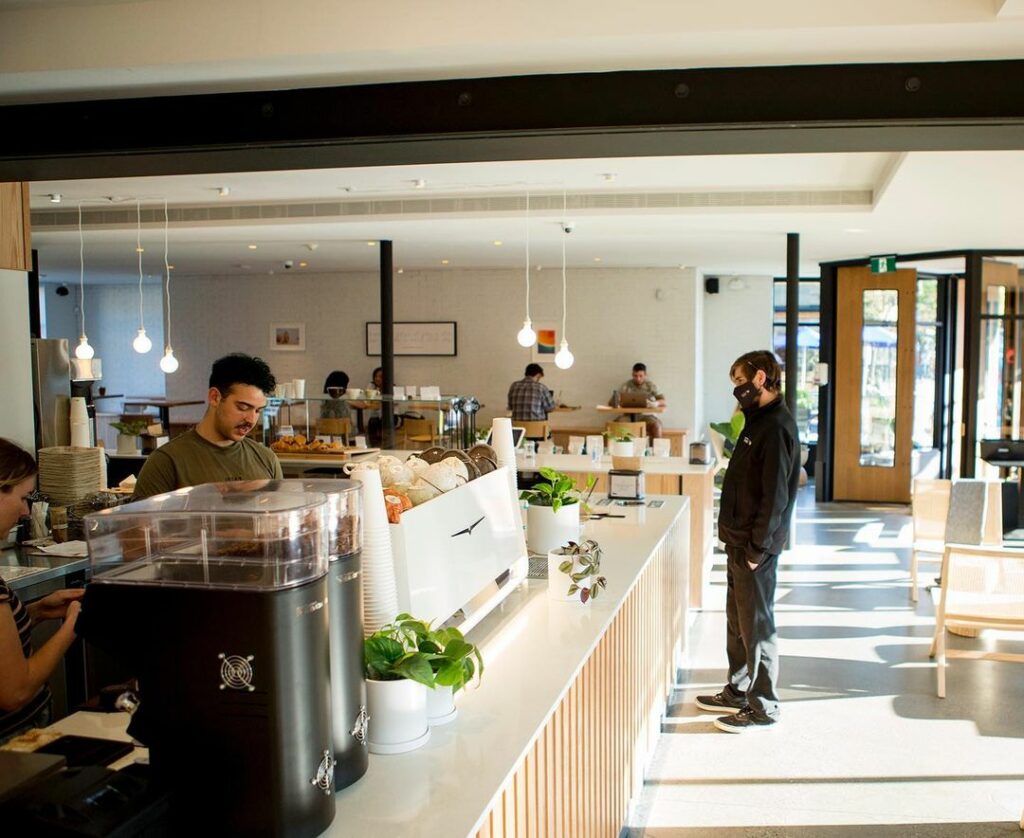 A man stands looking relaxed as a barista makes his coffee. It's a bright sunny day and the cafe is mostly black, white and wood.