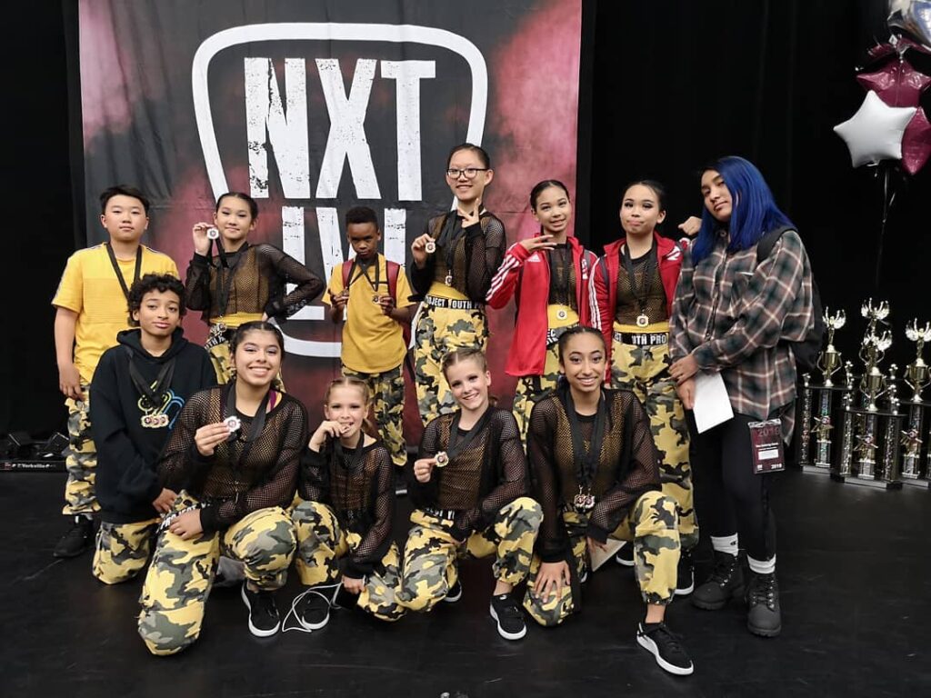 A group of young teen stand together posing in group looking at the camera holding dance competition medals. They have black tops and yellow camo pants.