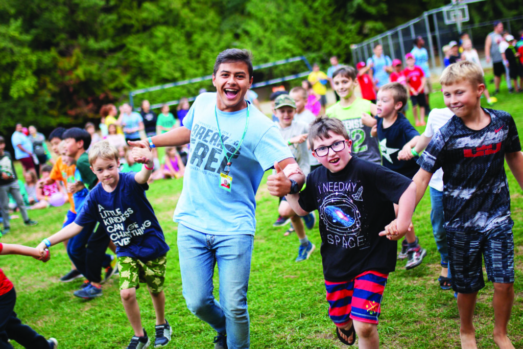 A camp director holds hands with three boys as they run smiling into the camera.
