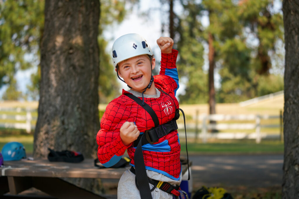 A boy is in climbing gear, he smiles and raises a joyful fist.