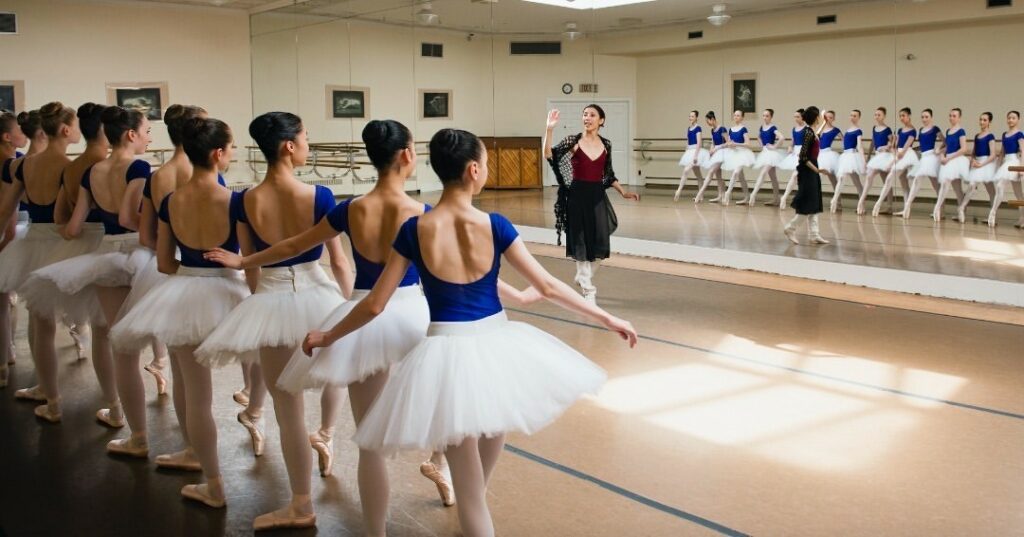 A long line of teen ballerinas in blue tops and white tutus pose in front of a dance instructor. The dance instructor stands in front of a mirror with her hand up, she is dressed in a maroon top and black skirt.