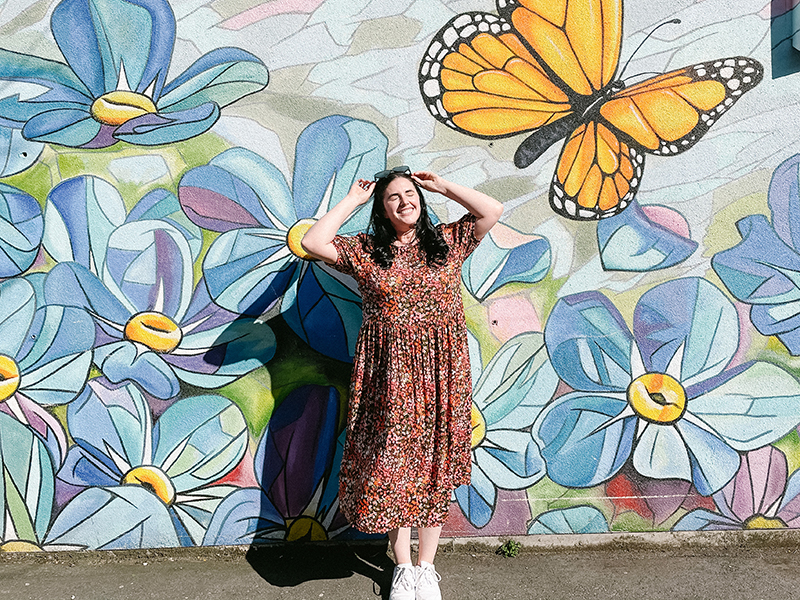 A woman stands with her face towards the sun with her eyes closed because it so bright. She stands in front of a mural with flowers and a butterfly.