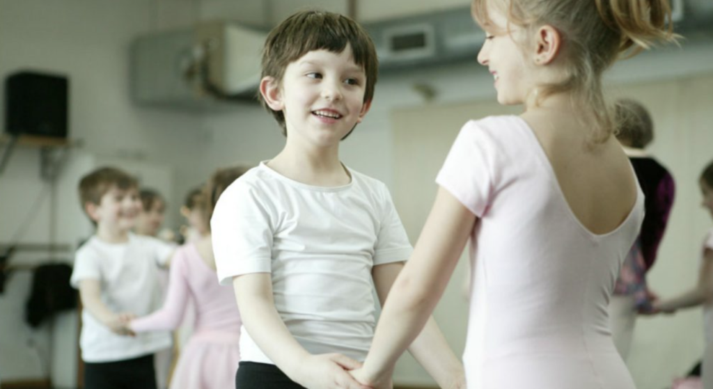 A young girl and boy hold hands and smile at each other with other children doing the same in the background. The children are dresses in traditional dance ballet clothes. The girl in a pink unitard with white tights and the boy in a white shirt and black leotards.