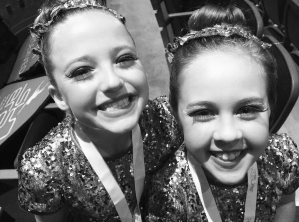 A close up photo of two young girl smiling into the camera. they have glittery tops and braided hair.