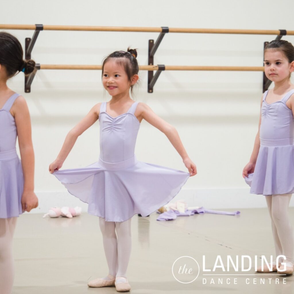 Three young dancer stand looking to the left of the camera. the first girl is cut off from the photo, the second girl holds her shirt out and smiles, the third girl looks serious.