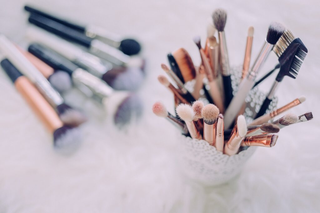 A lacy cup holds multiple make up brushes including blush brushes and eyebrow combs. On the ground blurred in the background are more make up brushes.