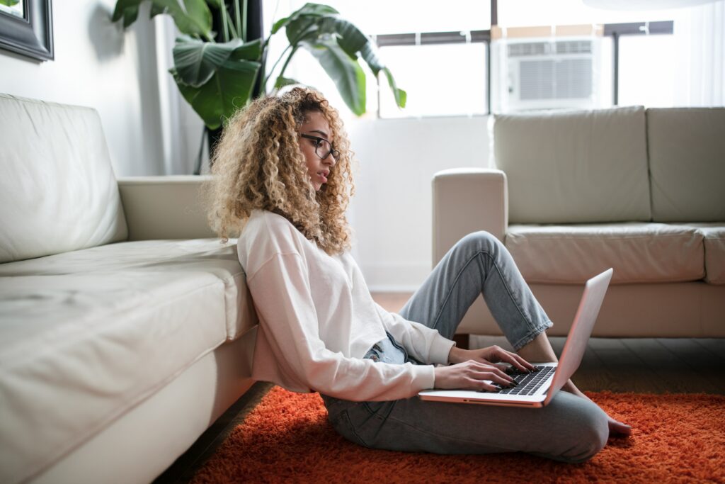 A woman reclines against a white couch on a burnt orange fuzzy carpet. Her lap top is open and she is looking down through her black rimmed glasses typing.
