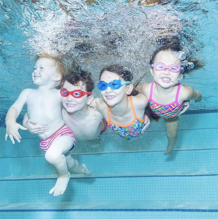 Four children under water smiling with bubbles coming out of their mouth. Three children wear swim goggles in red, blue and pink, and one baby with his eyes open and no goggles.