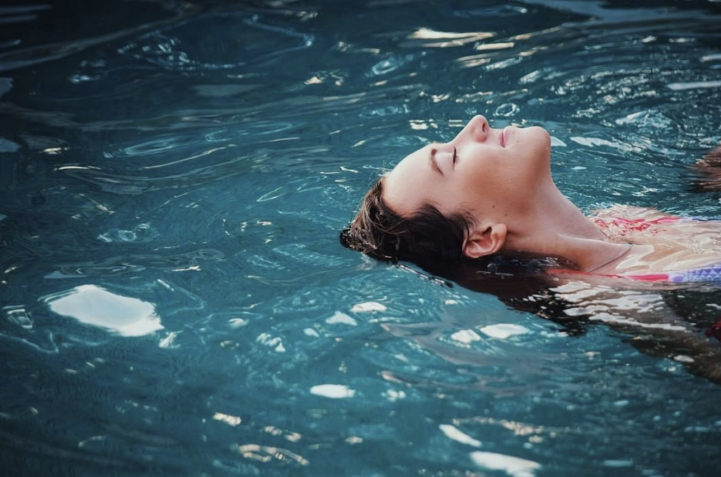 An older child floats on top of swimming pool water. She is smiling and you can see her from the shoulder up.