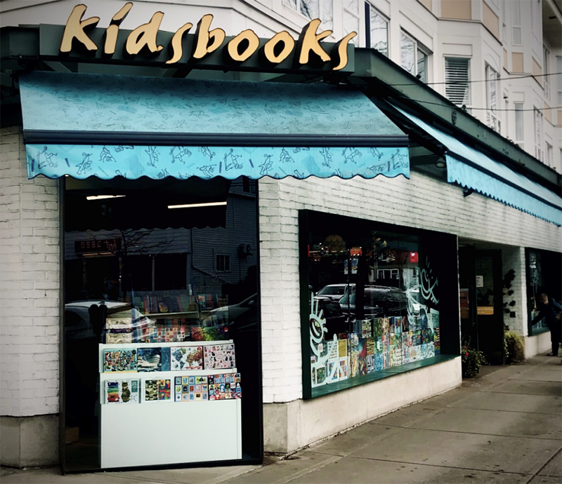 A photo of a corner of a building. With blue awning edges the window and a yellow sign reads Kidsbooks.