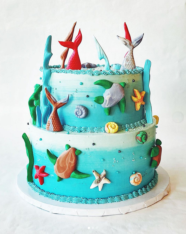A two layered blue cake decorated with sea creatures topped with mermaid tales.