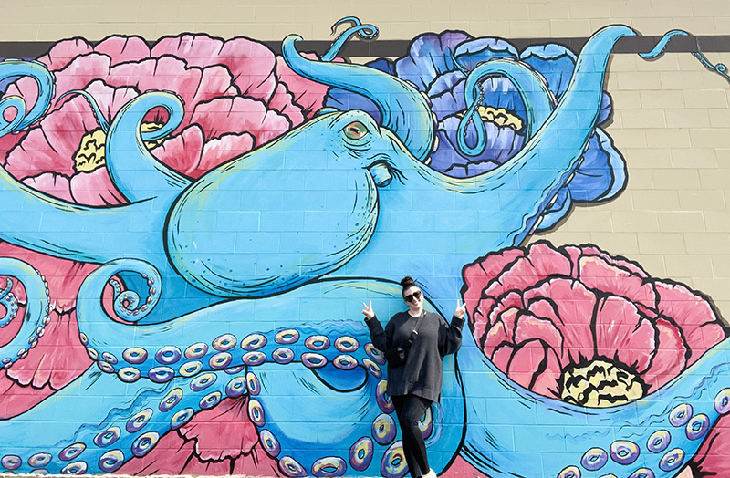 A woman in all black holds up two peace signs in front of a giant blue octopus.