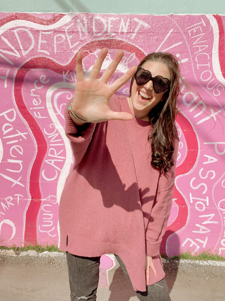 A woman in a pink shirt and grey pants holds up a high five in front of a pink mural with white words.