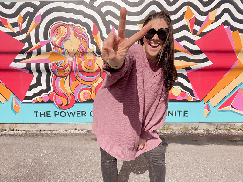 A woman in a pink shirt and grey pants with black heart shaped sun glasses gives a peace sign in front of a brightly coloured all that says The Power of Women is Infinite.
