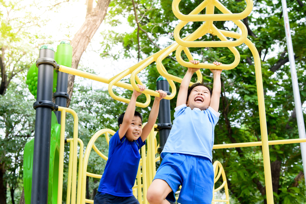 Two young children laugh as they hang from yellow monkey bars.