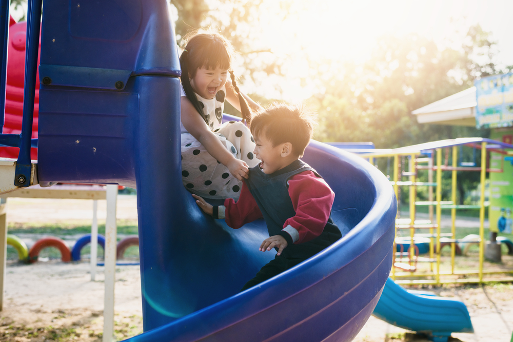 A little girl and boy go down a blue slide. The little girl is holding onto the boys shirt, both are laughing.