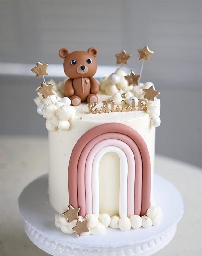 A tall white cake with an ombre rainbow topped with a brown bear and gold letters that read Kallie.