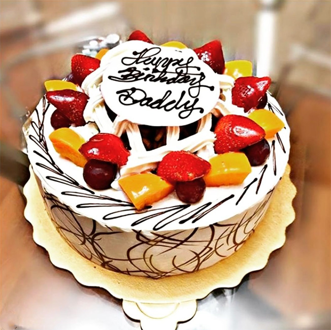 A white round cake topped with strawberries, mango, and grapes. On the top is a sign that says Happy Birthday Daddy.