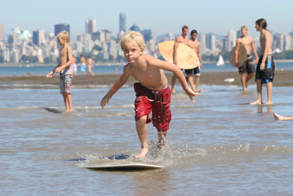 A little boy in red shorts has a look of concentration on his face as he jumps on a skimboard. Other boys are the background holding skimboards.