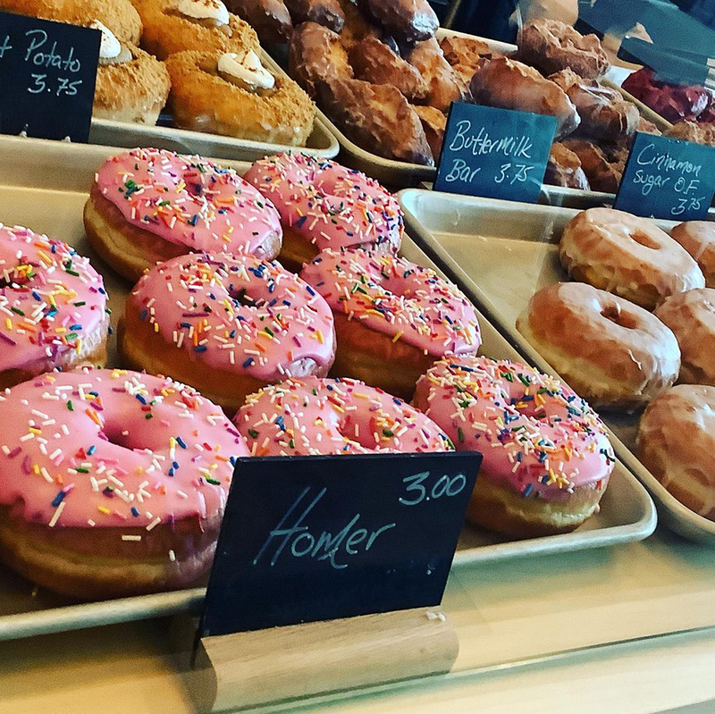 A close up of a donut shop window with a sign that says "homer". Its a pink donut with rainbow sprinkles.