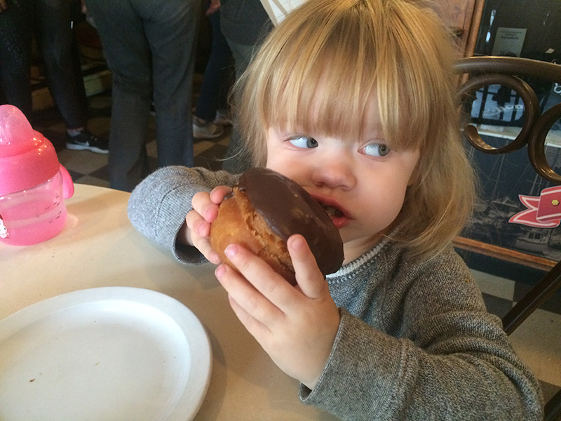 A little kid holds a chocolate covered donut to her mouth as she looks to the right of the photo.