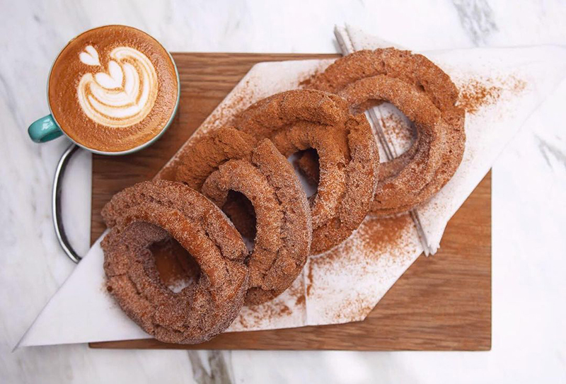 Four cinnamon covered donuts sit on a wooden trey by a cafe latte.