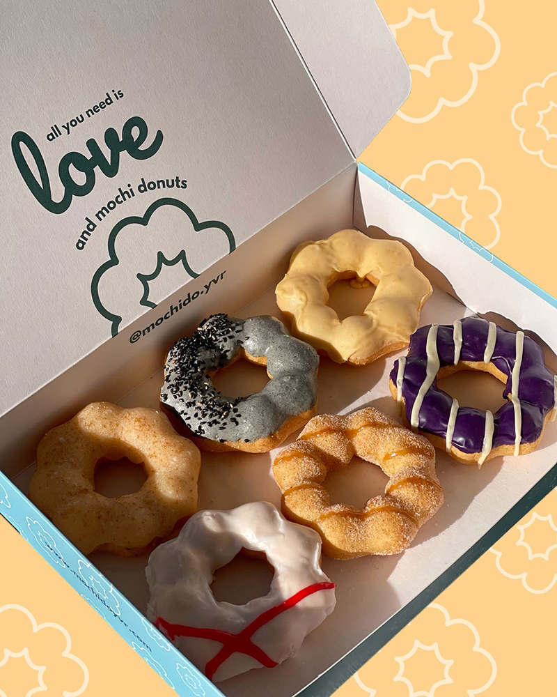 An open box of 6 donuts that are shaped like flowers. Two have orange icing, one grey with black sprinkles, one purple and one white.