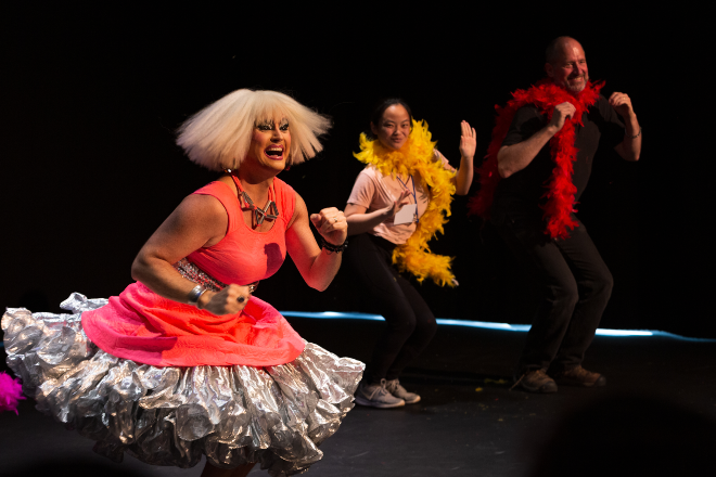 A woman in drag dances on a mostly black stage with a woman and a man in feather boas off to the side.