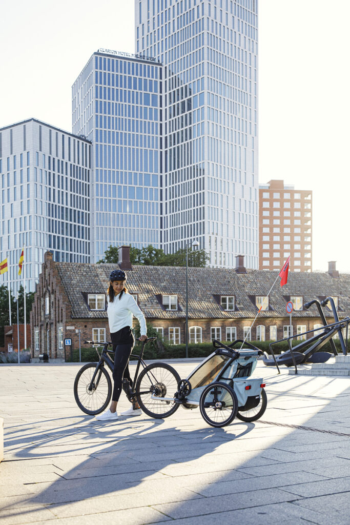 A woman in a courtyard stops on her bike and looks back at the child bike carrier attached to her black bike.