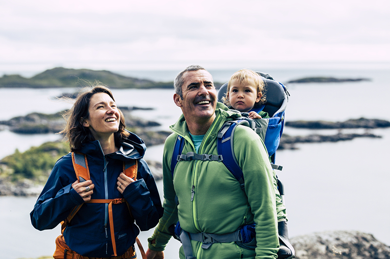 A man and a woman are on a hike smiling as they carry a baby in a child carrier backpack.