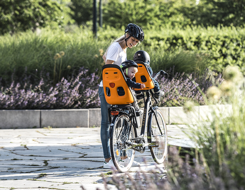 A woman looks back at two orange child bike seats carrying two small children.