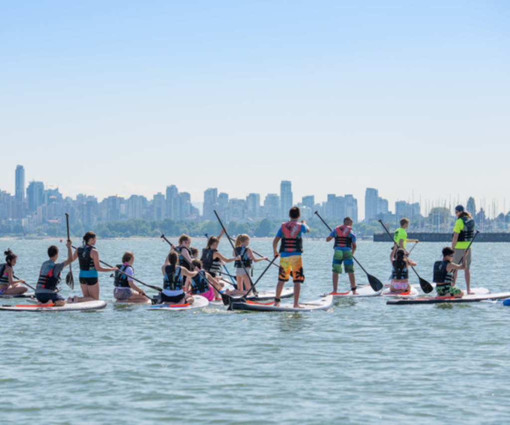 A large group of kids ride paddleboards with life vests on. Some stand while others sit.