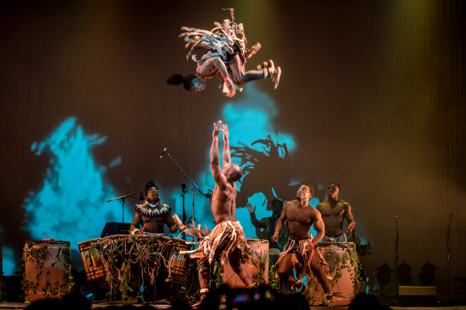 A man throws a woman high in the air, the other performers all look up.