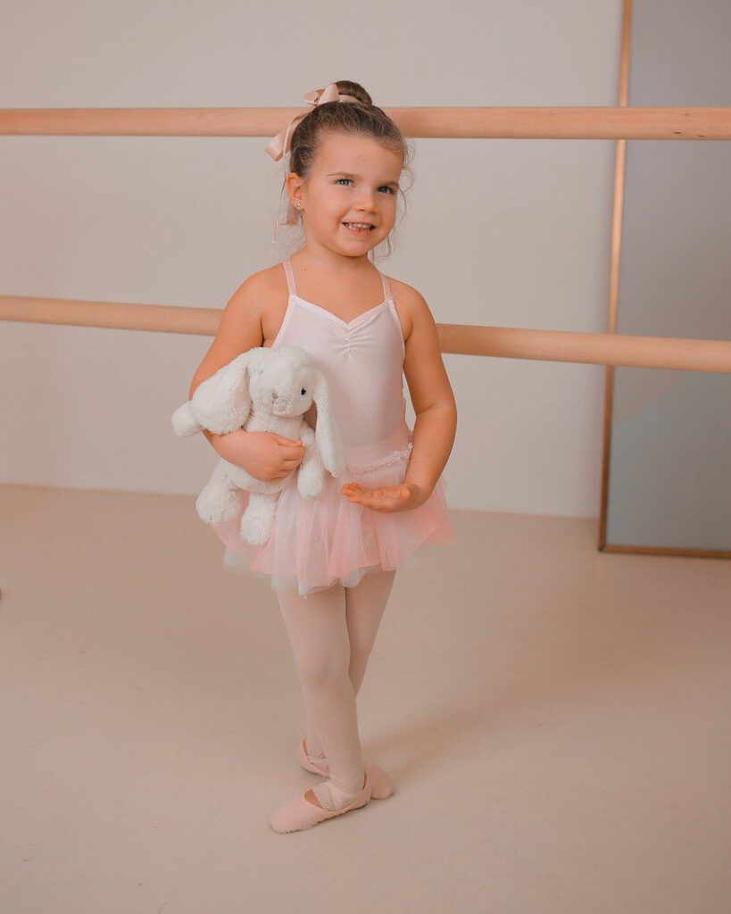 A little girl dressed in traditional ballet workout clothes stands posed with a stuffed bunny.