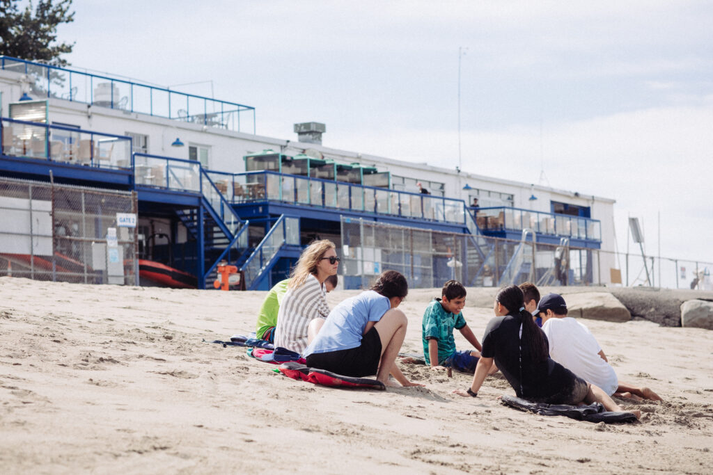 A group of lids and one adult sit in the sand in a circle on the beach.