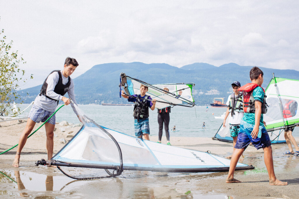 A group of boys in life vests carry windsurfing boards. One boy has a water hose and cleans his sail with it.