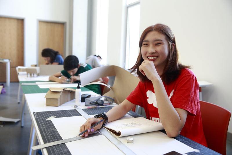 A smiling teen in red shirt looks into the camera with a pencil and ruler in hand at Emily Carr Teen Summer Camp.