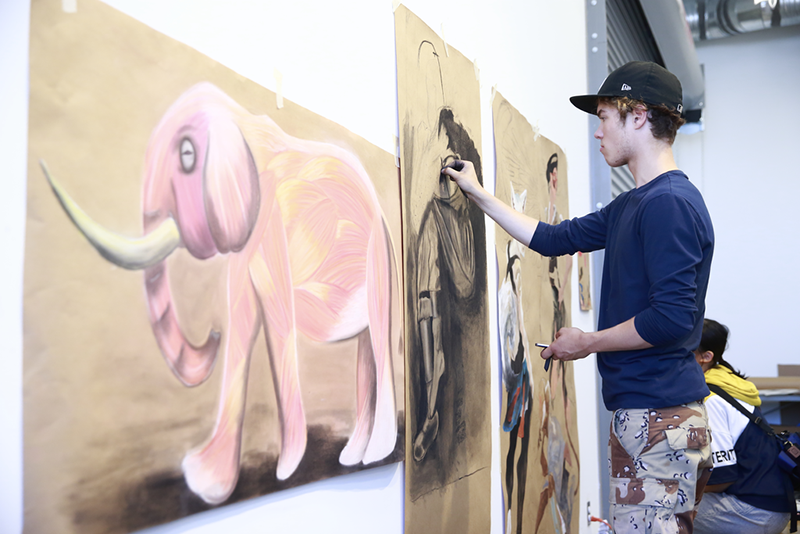 A teen boy stands up using charcoal to paint a figure on a large canvas at Emily Carr Teen Summer Camp.