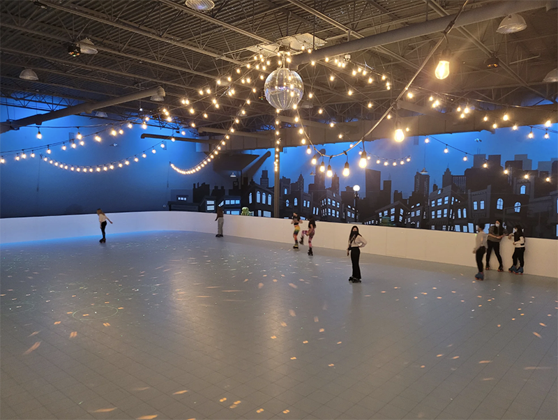 A picture of a large white skate rink with twinkle lights and a disco ball in the middle. 7 people skate in various places.
