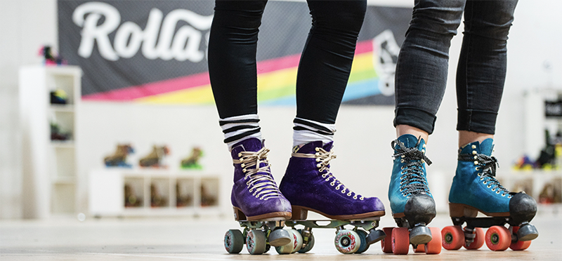 Picture of two people in black tights from the knee down wearing roller skates. One pair is purple and the other is teal.