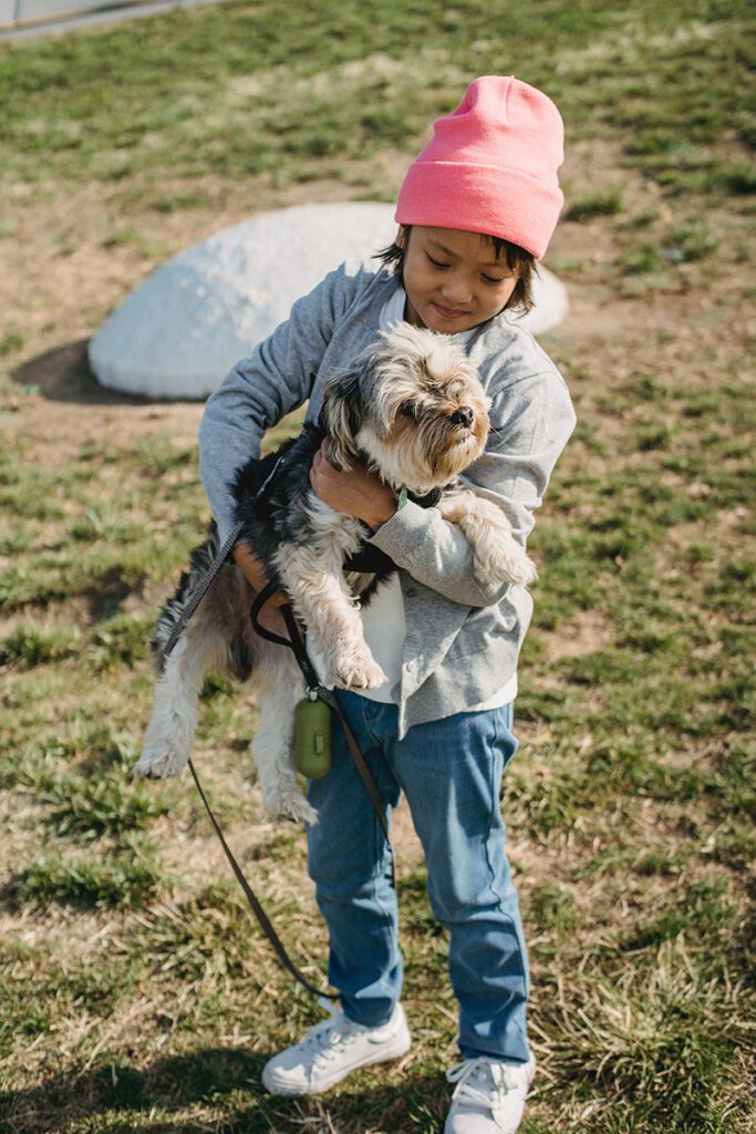 A little kid in a pink beanie holds a yorkie dog in his arms, the boy is smiling at the dog.
