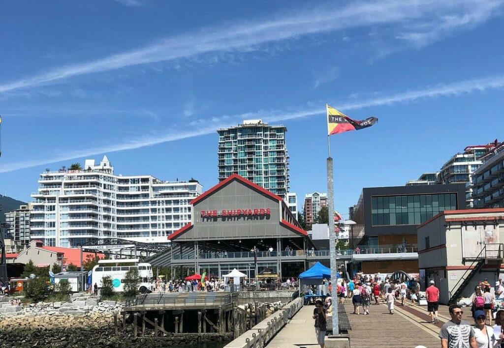 A photo of The Shipyards in North Vancouver. A building reads The Shipyards in red on a grey building with the city in the backdrop.