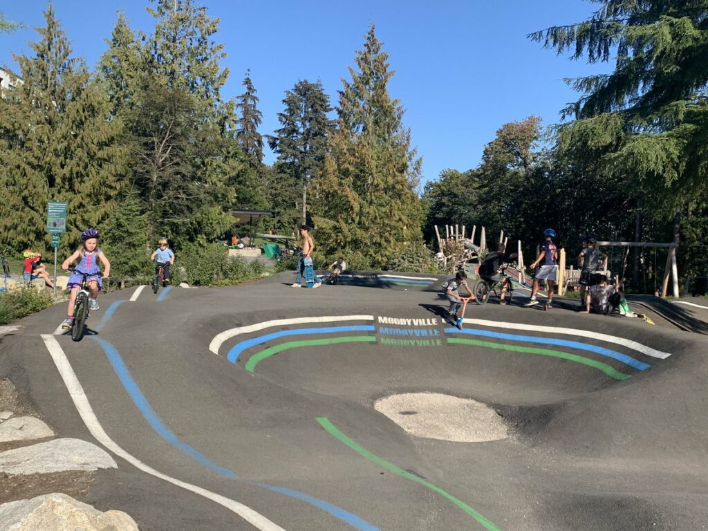 Various kids on bikes with helmets on ride in an asphalt bike bowl. The bowl has green and blue lines painted on it.