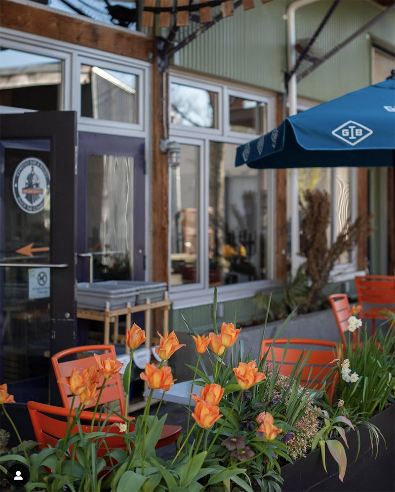 A sunny day photo of the outside of Off the Tracks on Granville island. A shop door is surrounded by orange tulips.