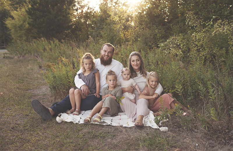 A dad, mom and 4 girls sit on a blanket in the tall grass all smiling and looking at the photo. Photo for VM Family Photographers.