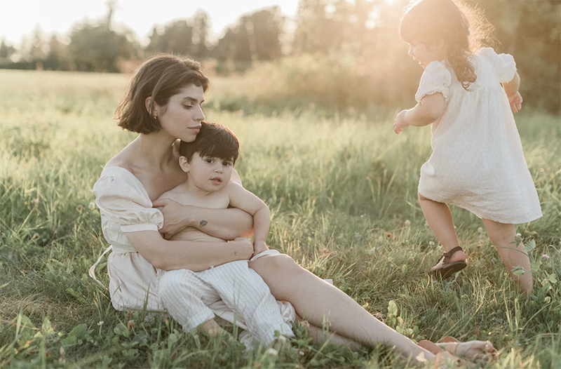 A mother closes her eyes as she holds a toddler with no shirt on, while a little girl dances in the grass. They all wear white on a grassy field. Photo for VM Family Photographers.